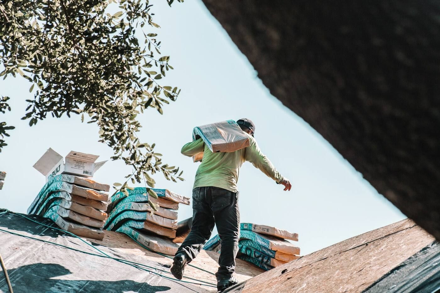 A roofer carrying a bundle of shingles across a stripped roof deck, with more bundles stacked behind him